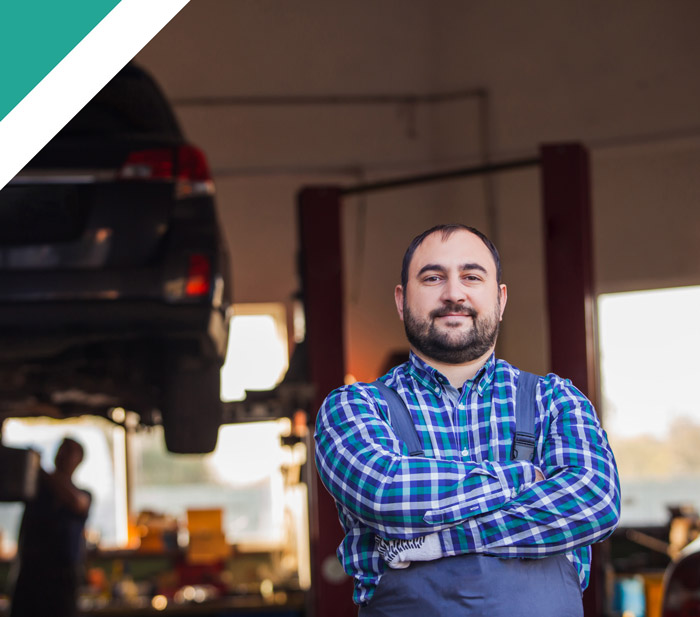 A confident mechanic in a workshop, wearing a plaid shirt and standing with arms crossed, with a car elevated on a lift in the background, showcasing automotive expertise.