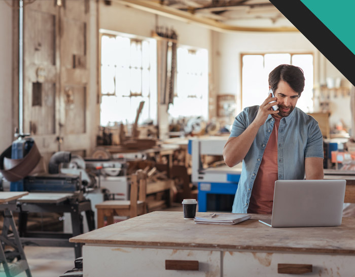 A man working in a workshop talking on the phone while using a laptop, surrounded by tools and wooden furniture.