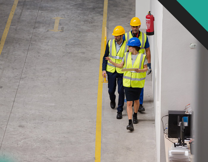 Three construction workers in safety vests and helmets walk through an industrial space, discussing plans while holding a clipboard.