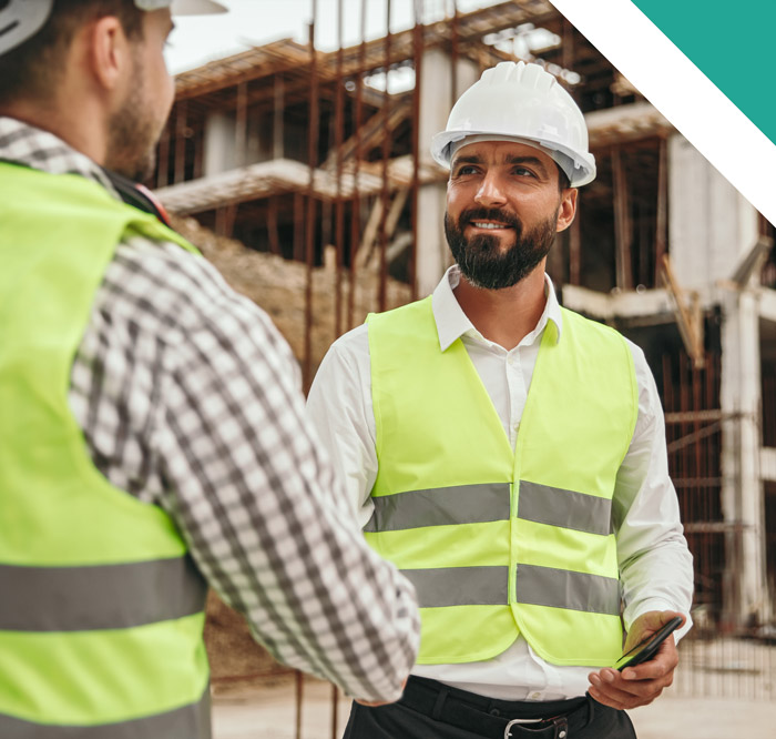 Two construction workers wearing protective gear and hard hats are conversing on a construction site, one holding a phone while smiling.