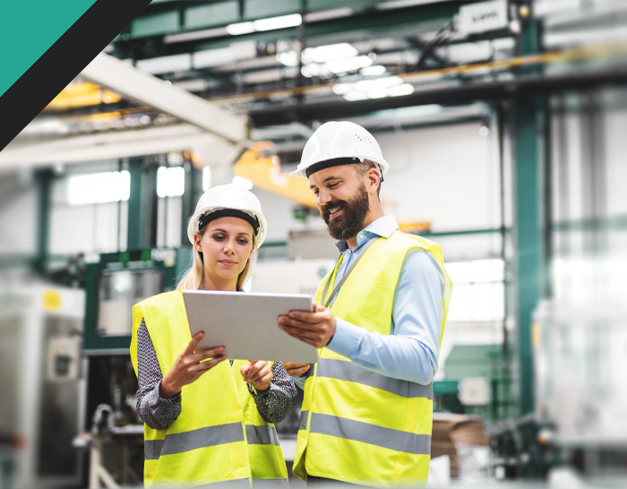 Two construction workers in hard hats and safety vests review a tablet in a factory setting.