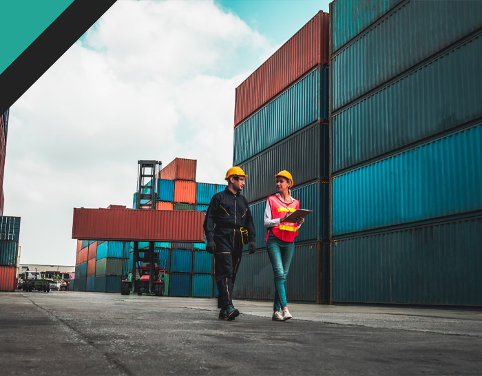 Two construction workers, one male and one female, are walking through a shipping yard filled with colorful containers, discussing plans and safety measures while wearing helmets and safety vests.