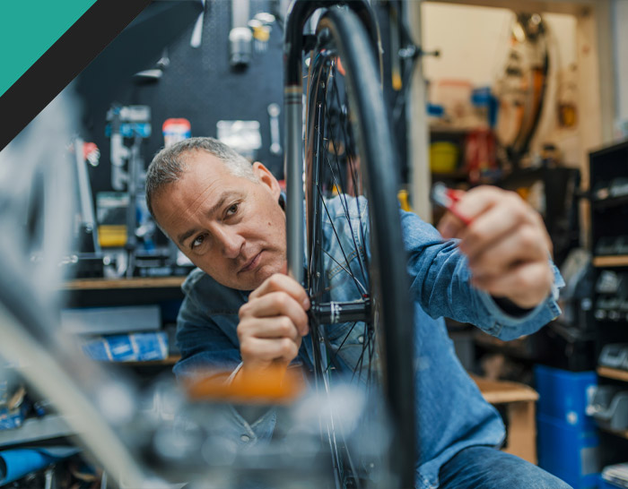 A man is repairing a bicycle wheel in a workshop, focused on adjusting components with tools at hand, surrounded by bike parts and equipment.