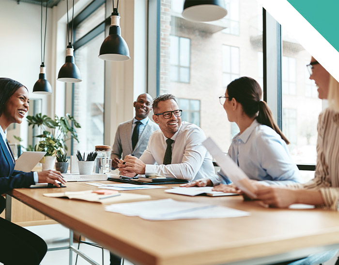 A diverse group of professionals engaging in a lively conversation around a conference table, with papers and notebooks on the table and large windows providing natural light.