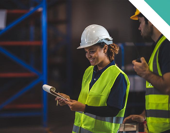 A smiling woman in a safety helmet and bright yellow vest is reviewing a clipboard, while a man in a yellow hard hat stands nearby, in a warehouse setting.