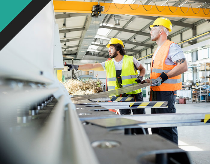 Two construction workers in safety gear, including helmets and reflective vests, operate machinery inside a large warehouse. One worker points at a control panel while the other holds a metal piece, demonstrating teamwork in an industrial setting.