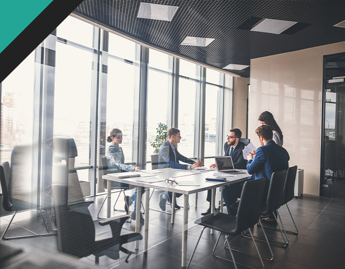 A modern office meeting with six professionals discussing around a conference table, large windows in the background allowing ample natural light, showcasing teamwork and collaboration.