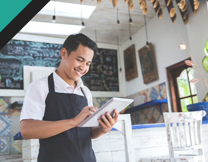 A smiling young man in an apron uses a tablet in a cafe, surrounded by a bright interior with decorative elements and chalkboards.