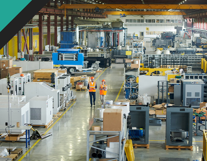 Two workers in safety gear walking through a large manufacturing facility filled with machinery and equipment, showcasing an industrial workspace.