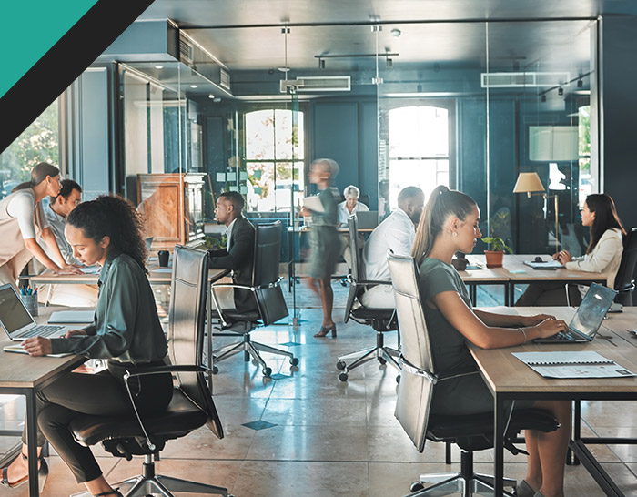 A modern office workspace featuring multiple professionals engaged in various activities, including working on laptops and discussing. The environment is bright and stylish, with glass walls and contemporary furniture, reflecting a productive and collaborative atmosphere.