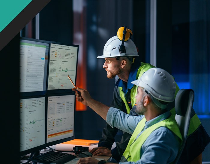 Two workers in safety helmets and vests analyze data on multiple computer monitors in a control room, discussing insights and operational performance.