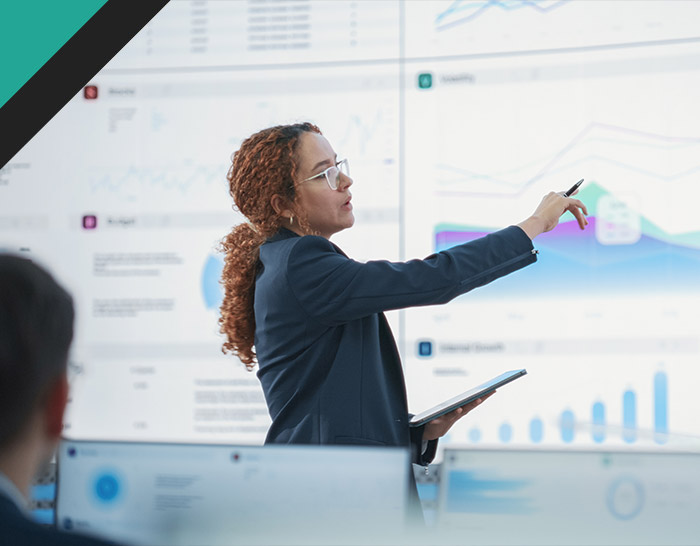 A woman in a business suit gestures while presenting data on a screen, holding a tablet in one hand. The presentation includes charts and graphs, illustrating analytical insights and business performance metrics.