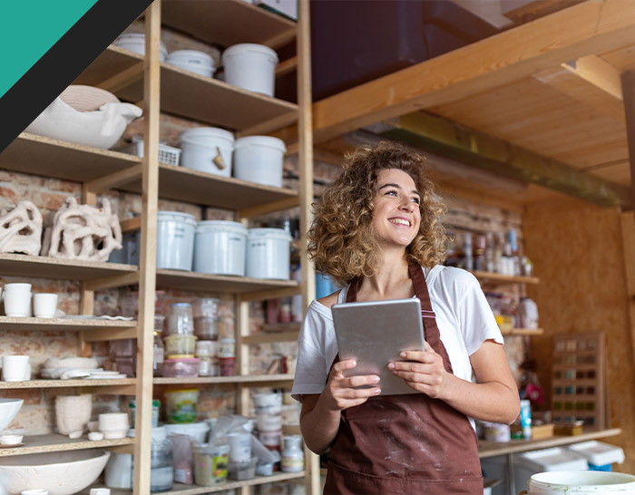 A smiling woman in an apron holding a tablet stands in a cozy workshop filled with shelves of art supplies and pottery.