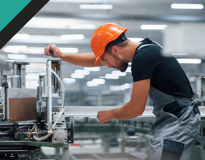 A male worker in an orange safety helmet and gray overalls is adjusting a machine in a bright industrial setting, focusing on his task with precision and care.