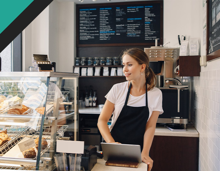 Barista working at a coffee shop counter, smiling while using a tablet, with pastries displayed in a glass case and a menu board in the background.