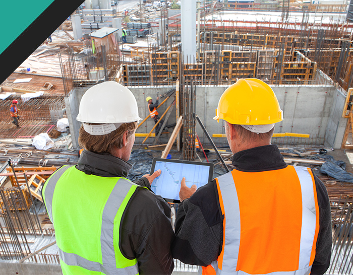 Two construction workers in safety gear reviewing data on a tablet at a construction site, surrounded by steel rebar and building materials, focusing on project management and coordination.