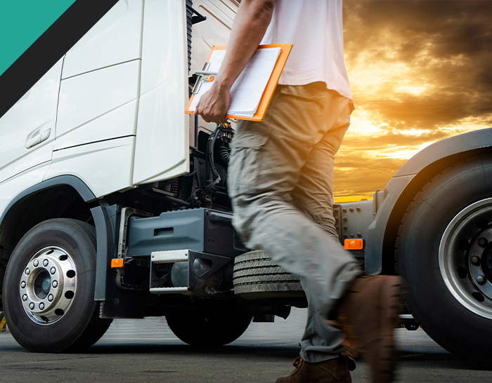 A technician walking beside a large truck, holding a clipboard, with a dramatic sunset in the background. Truck maintenance in progress.