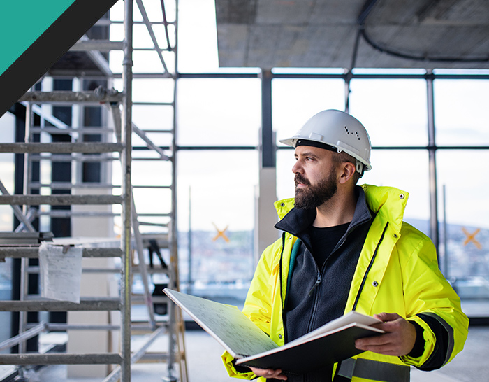 A construction worker wearing a hard hat and bright yellow safety jacket holds a clipboard while observing the worksite, with scaffolding and large windows in the background.