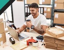 A man sitting at a table with a laptop, smiling while holding a coffee cup, surrounded by cardboard boxes and packing materials in a workspace.