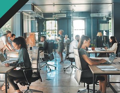 A modern office environment with people working at desks, some using laptops and others collaborating. The setting features large windows and a stylish decor typical of contemporary office designs.